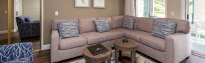 light gray sofa sectional with blue and white pillows, rug and chair. Coastal artwork and small coffee tables